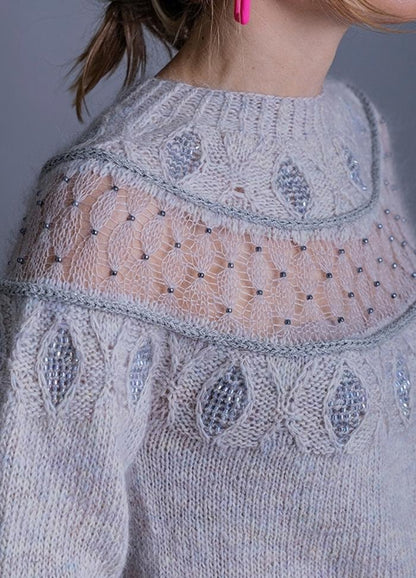 A close-up of a light gray knitted sweater with intricate lace detailing on the upper chest, featuring a geometric pattern. Bright pink earrings add contrast.