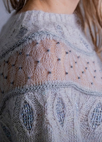 Close-up of a light grey knitted sweater with intricate lace patterns and tiny beads. The design features a delicate, airy texture, conveying elegance.