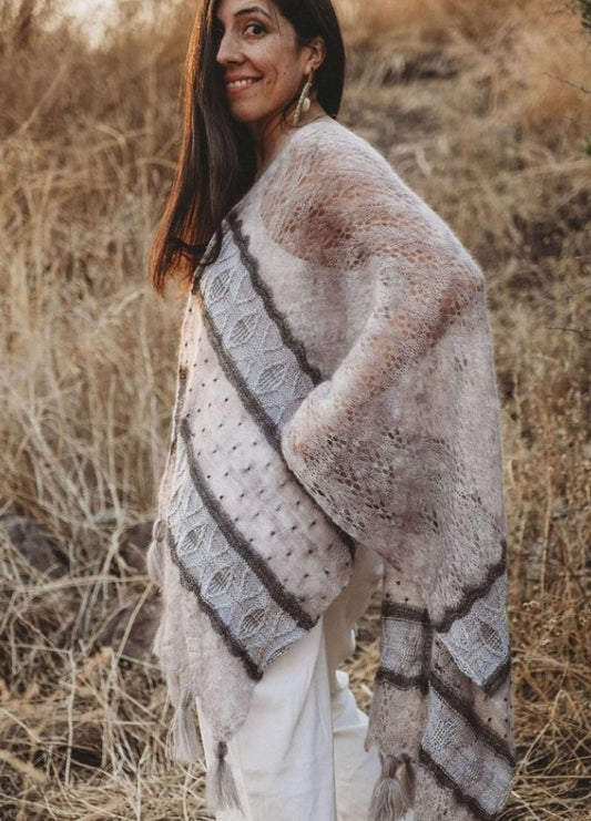 Woman outdoors in a grassy field wearing a patterned shawl with earthy tones. She smiles softly, evoking warmth and serenity in a natural setting.
