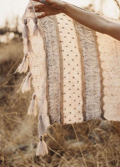 A hand holds a delicate, knitted fabric with intricate patterns and tassels, set against a blurred, sunlit, dry grass background, conveying warmth and artistry.