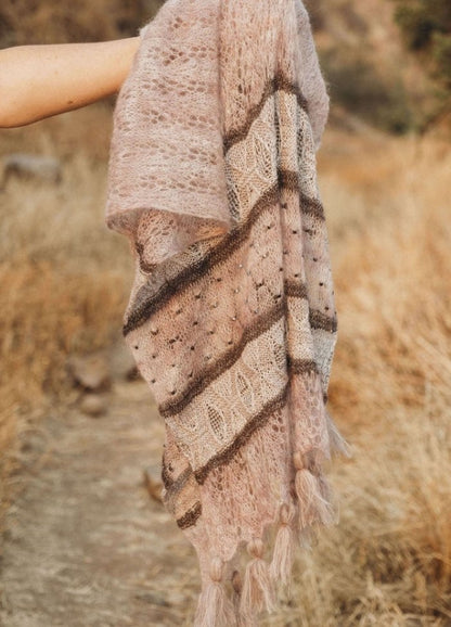 A hand holds a delicate, textured shawl with intricate patterns and tassels in earthy tones, set against a blurred backdrop of dry, golden grass.