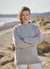 A woman in a gray sweater stands on a sandy beach with arms crossed. The background is a serene, blurred shoreline and gentle dunes under a clear sky.