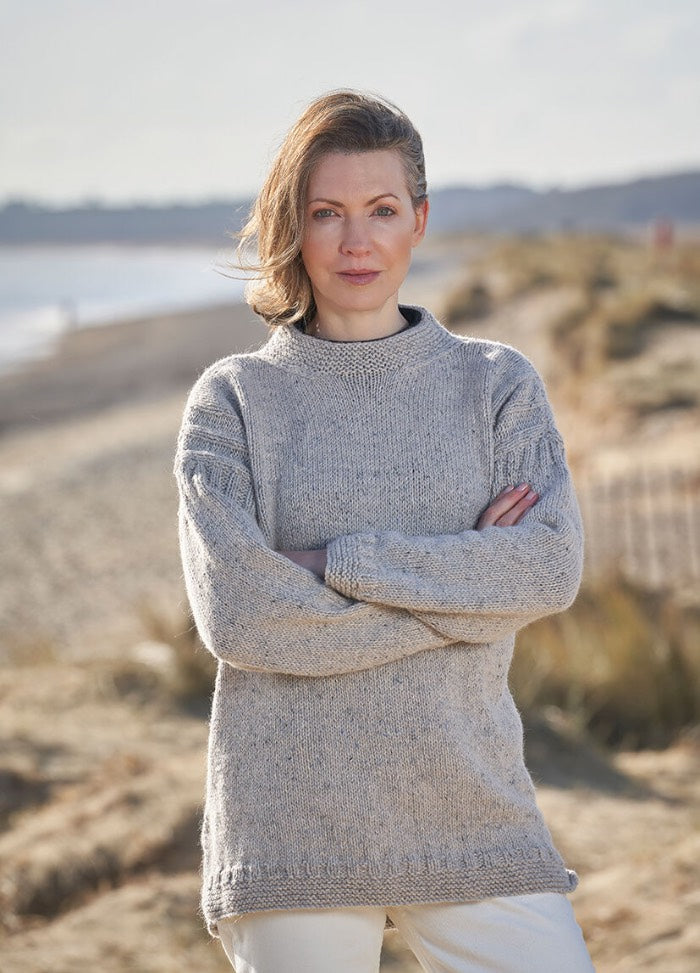 A woman in a gray sweater stands on a sandy beach with arms crossed. The background is a serene, blurred shoreline and gentle dunes under a clear sky.
