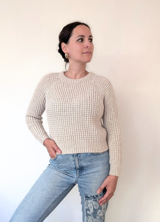 A woman in a beige knit sweater and blue jeans poses confidently against a plain background. She looks to the side, exuding a calm and thoughtful demeanor.