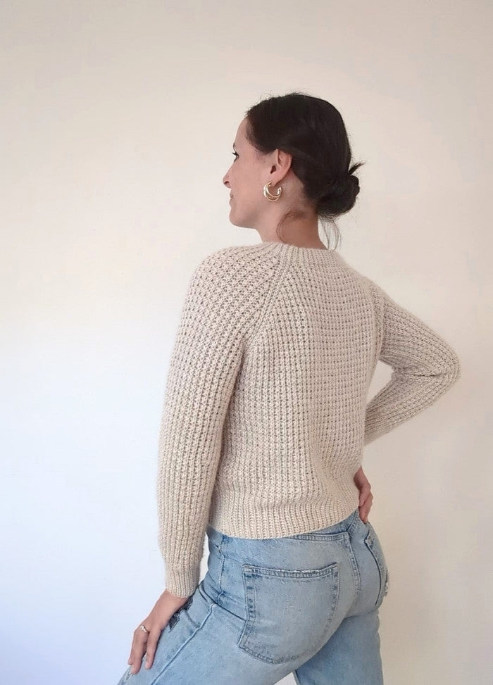 Woman in cream knit sweater and light blue jeans, turned sideways, smiling against a plain background. Gold hoop earrings add a stylish touch.