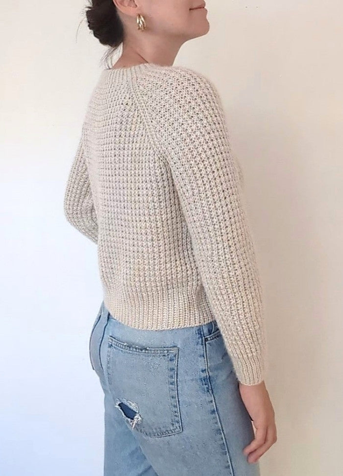 Woman in a textured cream sweater and light blue jeans with a pocket tear, standing sideways. Her hair is in a bun, and she wears gold hoop earrings.