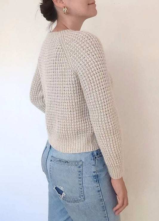 Woman in a textured cream sweater and light blue jeans with a pocket tear, standing sideways. Her hair is in a bun, and she wears gold hoop earrings.