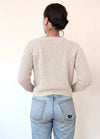 A person with their back to the camera is wearing a cream knit sweater and light blue jeans with a small tear. Their hair is in a bun, and gold hoop earrings are visible.