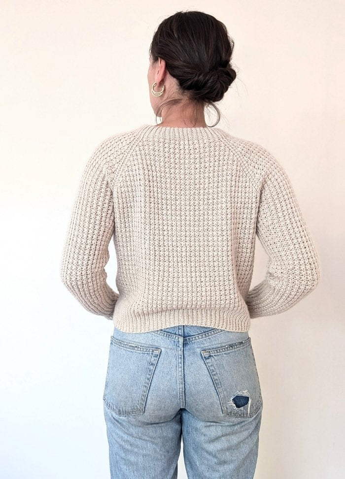 A person with their back to the camera is wearing a cream knit sweater and light blue jeans with a small tear. Their hair is in a bun, and gold hoop earrings are visible.
