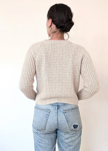 A person with their back to the camera is wearing a cream knit sweater and light blue jeans with a small tear. Their hair is in a bun, and gold hoop earrings are visible.