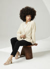 A woman with an afro sits on a wooden stool, wearing a cream cardigan, black pants, and white heels. The background is minimalist and soft-lit.