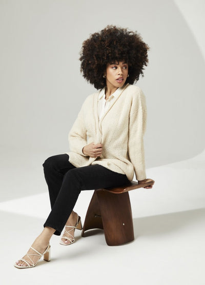 A woman with an afro sits on a wooden stool, wearing a cream cardigan, black pants, and white heels. The background is minimalist and soft-lit.