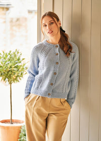 Woman in a light blue knitted sweater and tan pants leans against a beige wall. A potted plant with green leaves is in the background. Calm expression.