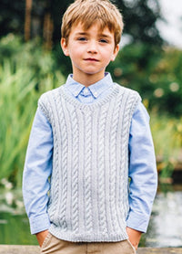 Edward Vest: Young boy standing outdoors in a park-like setting, wearing a grey hand-knitted cable-knit vest over a light blue button-up shirt.