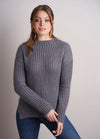 Woman in a cozy gray ribbed sweater and blue jeans poses against a light gray background, exuding a relaxed and content demeanor.