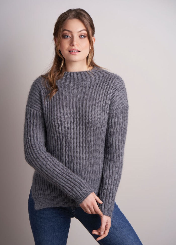 Woman in a cozy gray ribbed sweater and blue jeans poses against a light gray background, exuding a relaxed and content demeanor.