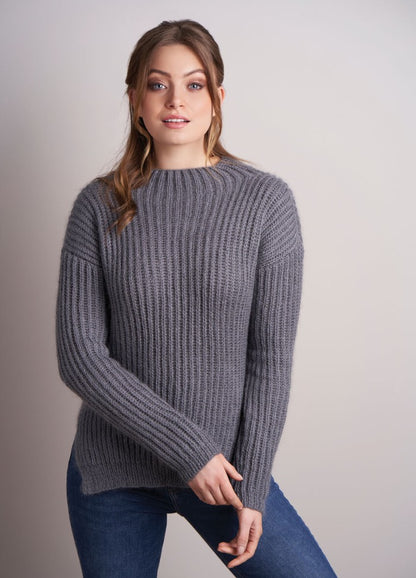 Woman in a cozy gray ribbed sweater and blue jeans poses against a light gray background, exuding a relaxed and content demeanor.