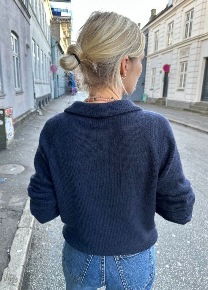 A person with blonde hair in a bun walks down a quiet street, wearing a navy Elisabeth Blouse and jeans. The scene is calm, with old buildings lining the street.