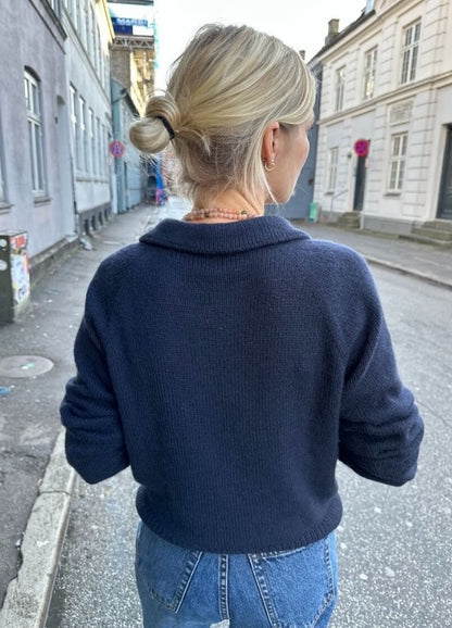 A person with blonde hair in a bun walks down a quiet street, wearing a navy Elisabeth Blouse and jeans. The scene is calm, with old buildings lining the street.