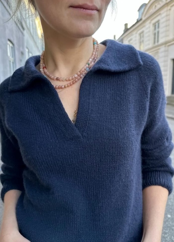 Woman wearing a navy Elisabeth Blouse with a V-neck, layered pink beaded necklaces, standing outdoors on a street with historic buildings. Casual and elegant.