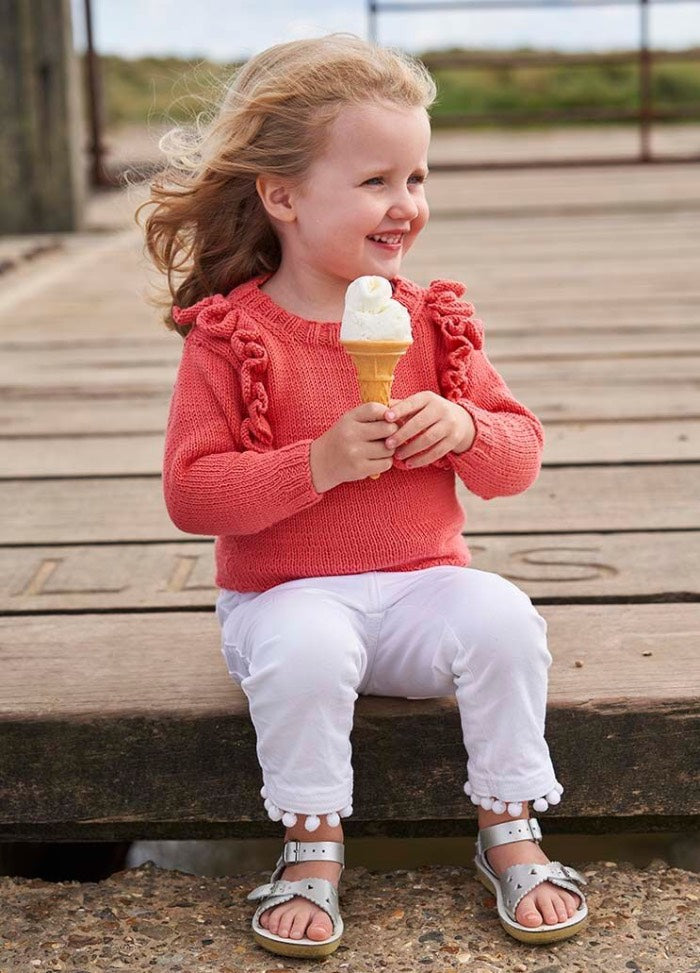 A smiling young girl with light brown hair sits on a wooden dock, holding an ice cream cone. She wears a coral sweater and white pants, exuding a joyful vibe.