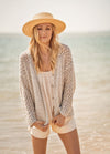 A woman with long blonde hair in a straw hat and crochet cardigan smiles gently at the beach. The background shows a calm sea under a clear sky.