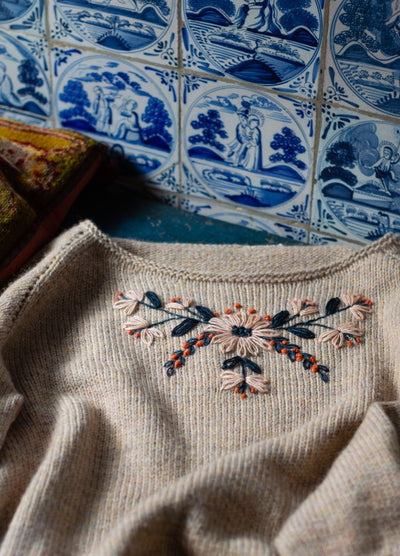 Higuchi san: black embroidery of stylized plants including tulip, sunflower, and budding branch on beige knit sweater.