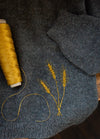 Rye: three yellow rye stalks embroidered on gray knit fabric beside matching thread spool.