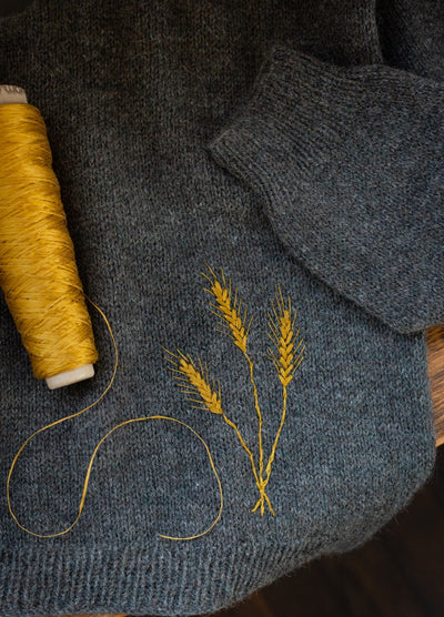 Rye: three yellow rye stalks embroidered on gray knit fabric beside matching thread spool.