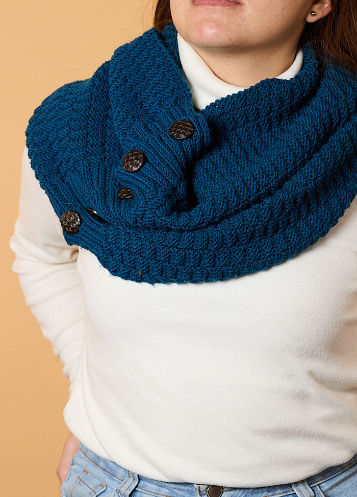 A woman in a white sweater wears a textured blue knit scarf with dark buttons. Set against a light brown background, the tone is cozy and stylish.