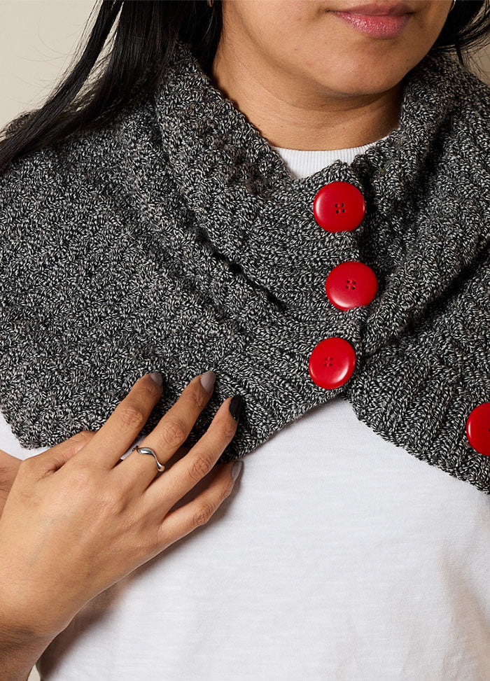 A woman wears a gray knitted scarf with three large red buttons. Her hand, with painted nails and a ring, touches the scarf. She wears a white top.