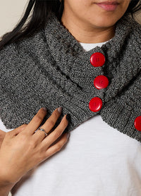 A woman wears a gray knitted scarf with three large red buttons. Her hand, with painted nails and a ring, touches the scarf. She wears a white top.