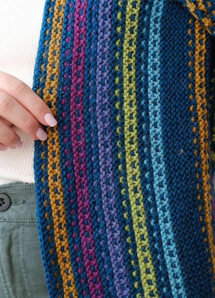 Close-up of Esme’s Sister Shawl with colorful stripes in blue, green, purple, pink, and mustard, held by hand over white top and green pants.