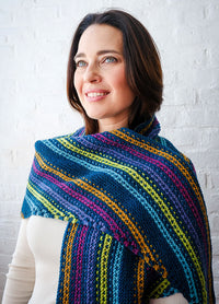 Person wearing Esme’s Sister Shawl with horizontal stripes in blue, green, yellow, pink, and purple, standing against white brick wall.
