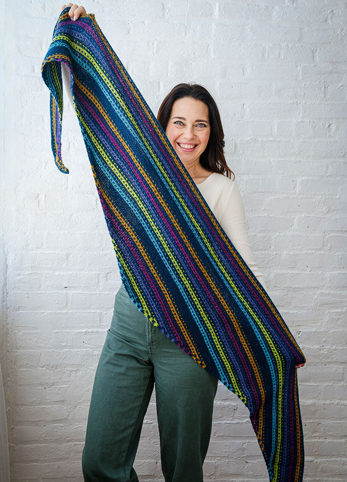 Person holding up Esme’s Sister Shawl with colorful horizontal stripes in blue, green, yellow, purple, and pink, against white brick wall.