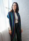 Person wearing Esme’s Sister Shawl with vertical stripes in blue, purple, green, and yellow, standing near window with white brick wall and sheer curtains.