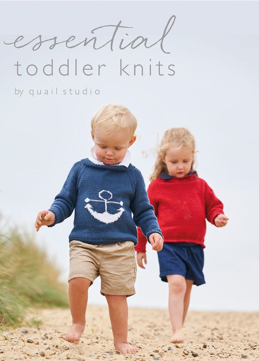 Two toddlers walking on a pebbly beach, wearing knitted sweaters; the boy in blue with an anchor design, the girl in red. Sky is overcast, evoking calm.