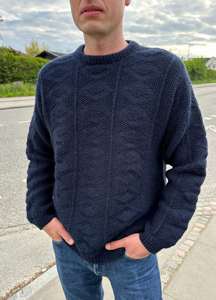 Man wearing the Esther Sweater in dark blue knit with textured pattern, round neckline, and long sleeves, styled with blue jeans in an outdoor setting.