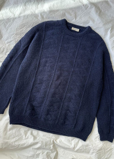 Flat lay of the Esther Sweater in navy blue textured knit with geometric pattern, round neckline, and ribbed cuffs, displayed on a white fabric background.
