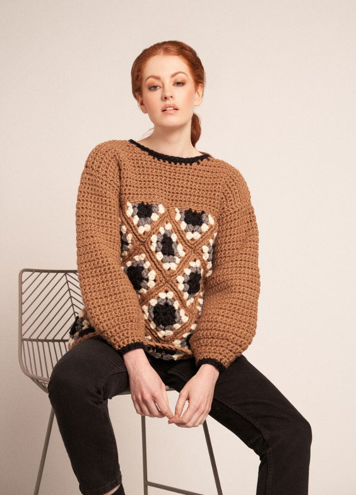 A woman sits on a chair, wearing a cozy brown and black crochet sweater with a textured pattern and a relaxed fit.