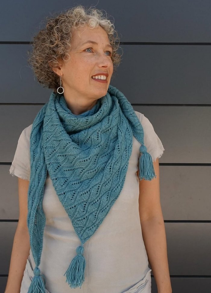 Woman with curly hair wearing a textured teal scarf with tassels, looking to the side. The background is a dark grey wall, and she appears thoughtful.