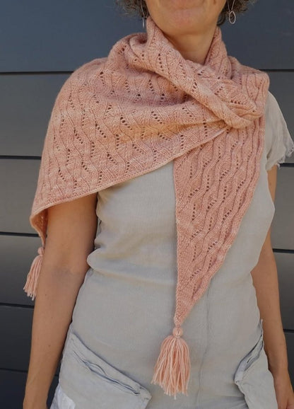 A person wearing a light pink, textured, knit shawl with tassels, draped over a beige top. The shawl has a cozy and elegant appearance.