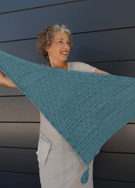 A smiling woman holds out a turquoise knitted shawl with a tassel, standing against a dark paneled background. She wears a light grey dress.