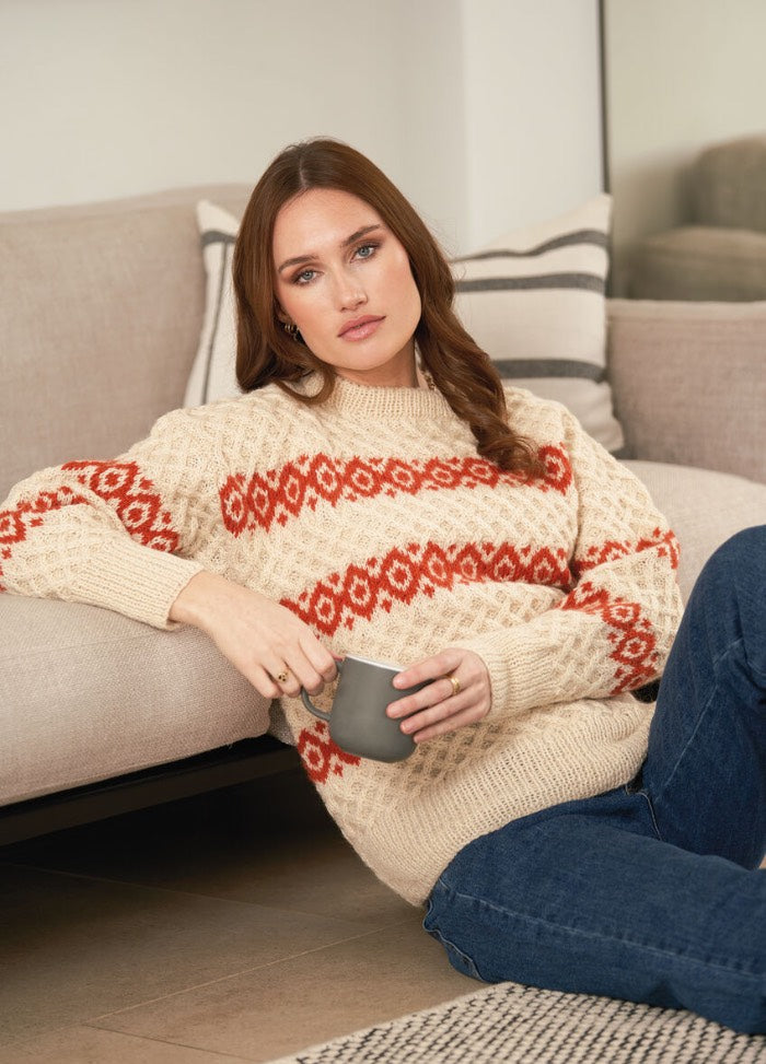 Woman lounging on a beige sofa, wearing a cream sweater with red patterns and blue jeans. She holds a mug, exuding a relaxed and cozy vibe.