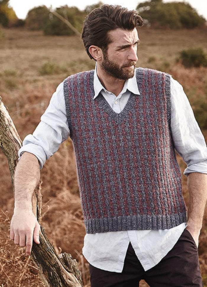 Person wearing the Faroe Vest, a striped knit design in grey and red with V-neck, layered over a light button-up shirt. Photographed outdoors with dry grass and bushes.