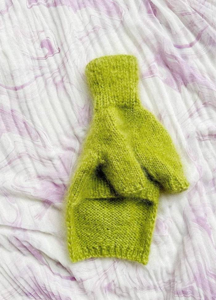 A single, bright green knitted dog jumper lies on a textured, white and pale purple marbled fabric. The scene feels cozy and soothing.