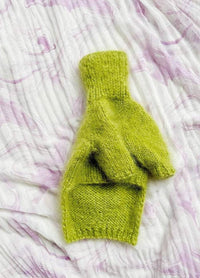 A single, bright green knitted dog jumper lies on a textured, white and pale purple marbled fabric. The scene feels cozy and soothing.