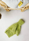 A fluffy green dog jumper lies on a light surface next to a wicker chair with a knitted blanket. A yellow "Kanto" tag is visible, creating a cozy scene.