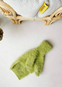 A fluffy green dog jumper lies on a light surface next to a wicker chair with a knitted blanket. A yellow "Kanto" tag is visible, creating a cozy scene.