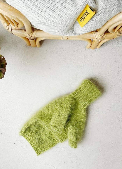 A fluffy green dog jumper lies on a light surface next to a wicker chair with a knitted blanket. A yellow "Kanto" tag is visible, creating a cozy scene.